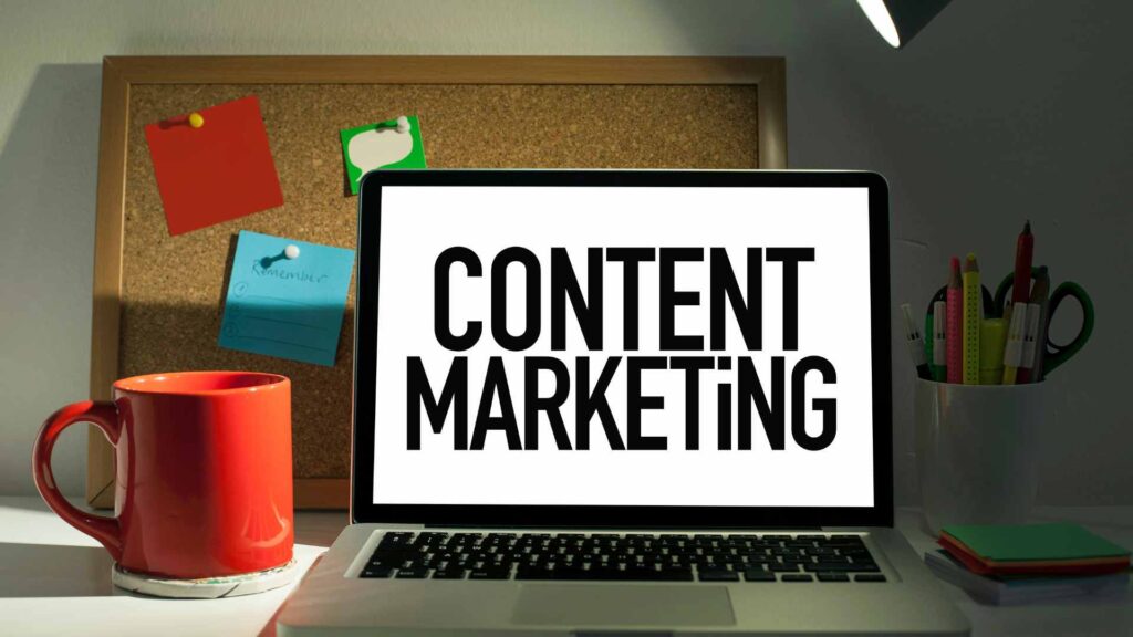 A laptop on a brightly lit desk displaying the words CONTENT MARKETING on the screen, next to a red coffee mug.